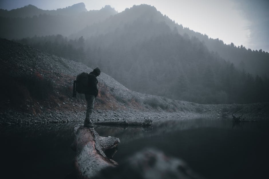 A solitary hiker traverses a log by a misty mountain lake, surrounded by nature's tranquility